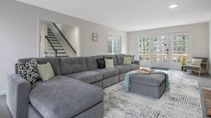 Een moderne woonkamer met een grijze modulaire bank, een klein poufje en een vloerkleed met een patroon. Er is een trap die naar een hoger niveau leidt, en een stoel bij een groot raam met uitzicht.