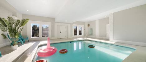 En indendørs swimmingpool med klart, blåt vand, omgivet af hvide vægge og døre. Der er farverige flyde ringe i poolen, og en potteplante er synlig på venstre side.