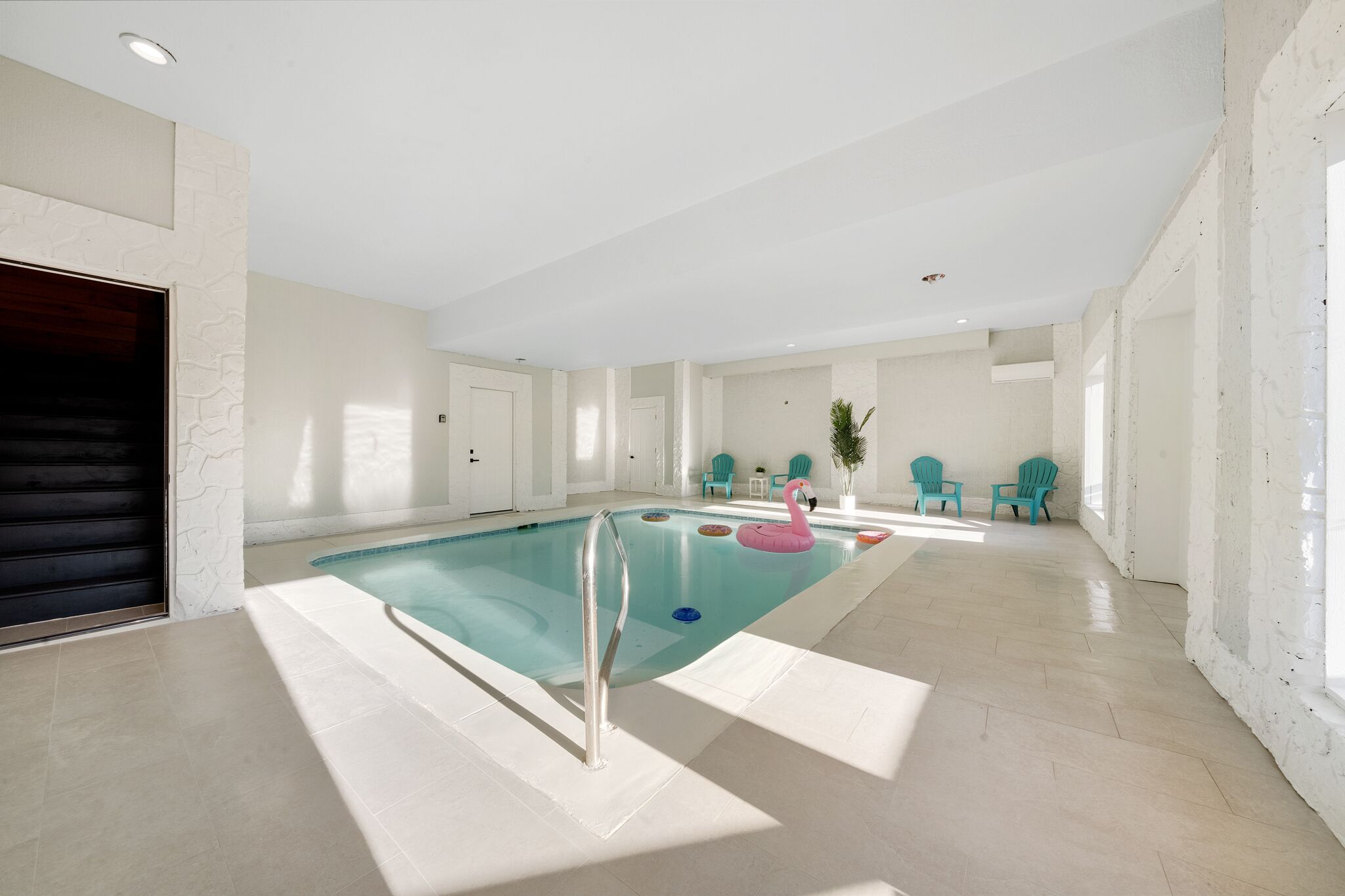 A spacious indoor swimming pool with a clear pool area, a pink flamingo float, and teal chairs in the background.