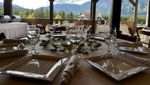 Une table dressée pour le dîner, avec une vue sur les montagnes, des poutres en bois et des guirlandes lumineuses.