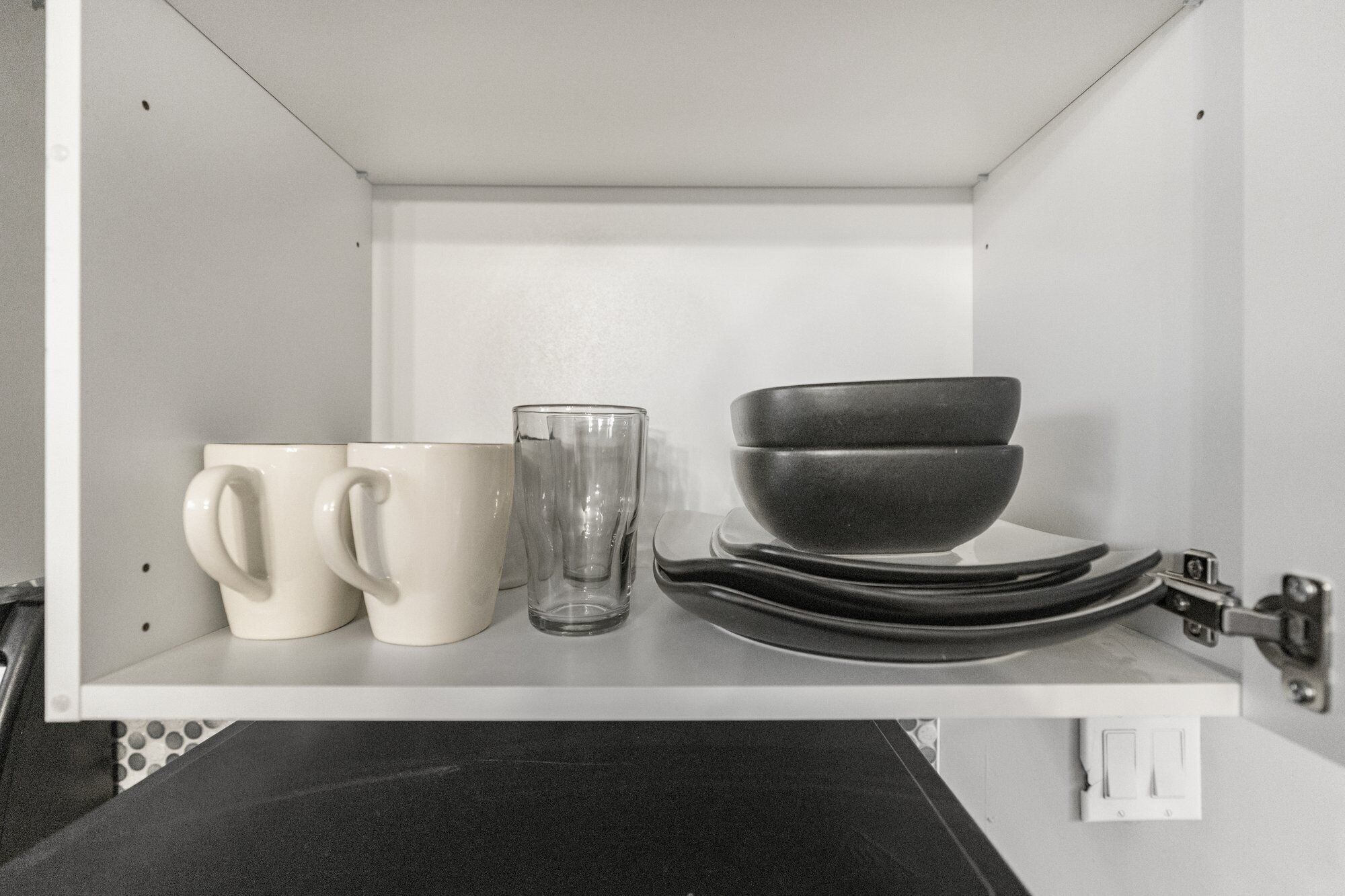 A kitchen shelf with white mugs, a clear glass, and stacked black bowls.