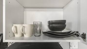 A kitchen shelf with white mugs, a clear glass, and stacked black bowls.