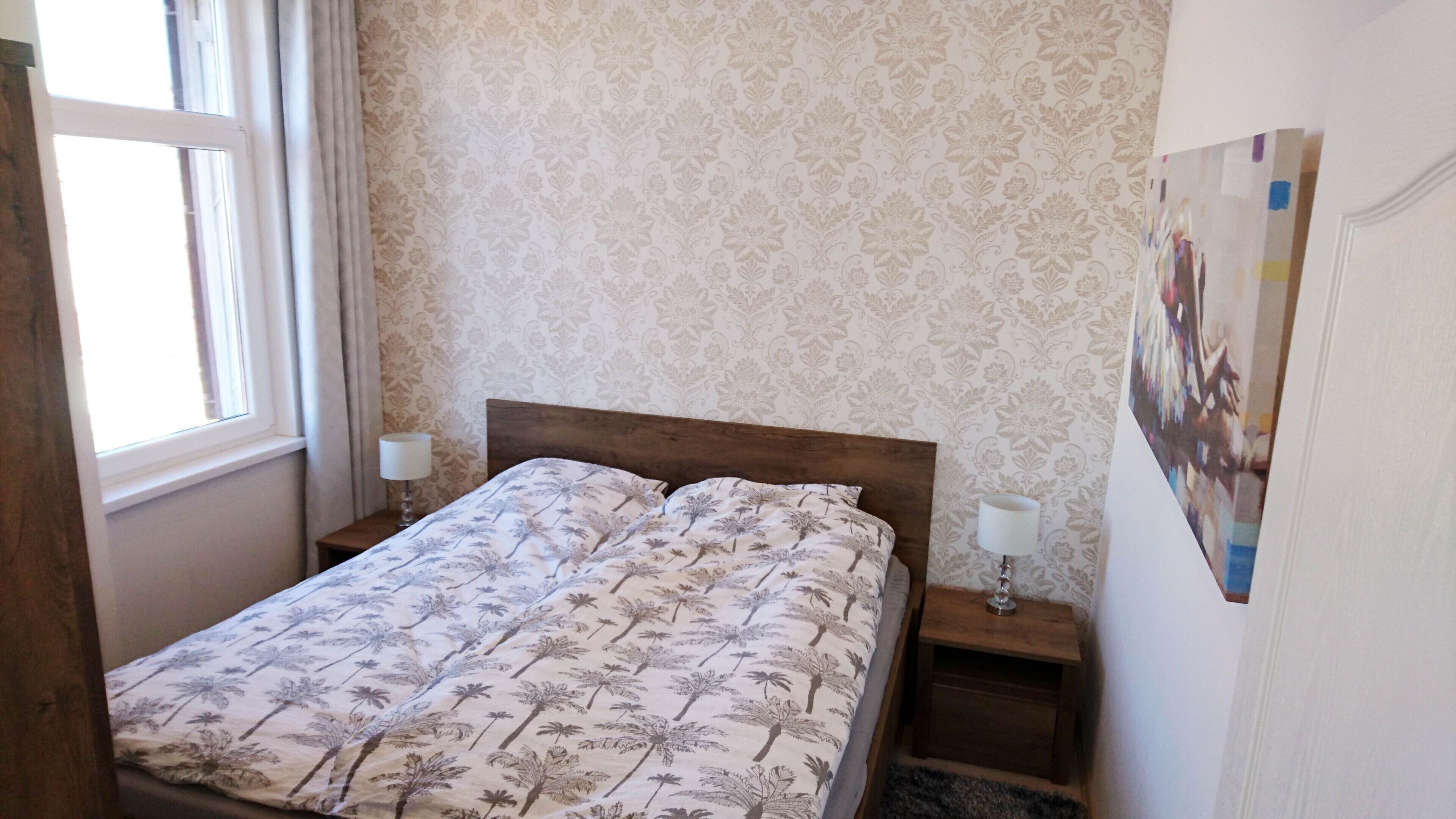 A neatly made bed with a floral bedspread, two bedside tables with lamps, a window with curtains, and a patterned wallpaper.