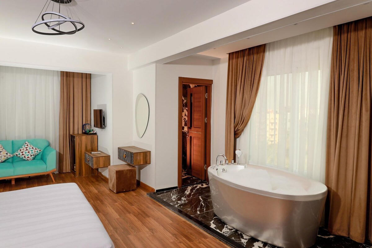 A modern hotel room with a large bed, a sitting area with a sofa, a wooden side table, a large bathtub, and a mirror on the wall.