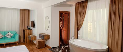 A modern hotel room with a large bed, a sitting area with a sofa, a wooden side table, a large bathtub, and a mirror on the wall.