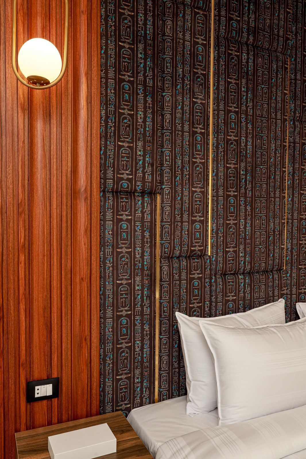 A hotel room with a wooden panel wall, a bed with white bedding, and a wall with hieroglyphic patterns.