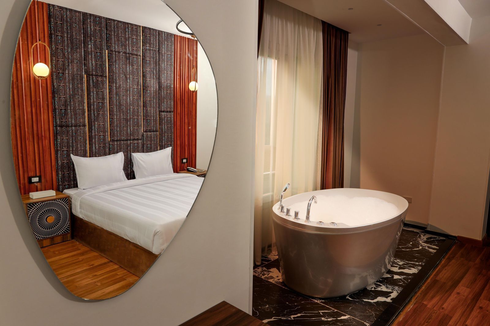 A hotel room with a large bed, a round mirror, a wooden headboard, a bedside table, and a large bathtub.