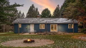 Une maison bleue avec un foyer au milieu de la cour, entourée d’arbres et d’un banc.