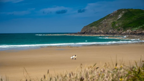 Una playa con dos personas llevando tablas de surf, un primer plano con hierba y una colina al fondo.