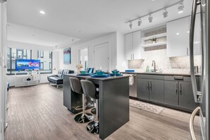 A modern kitchen with a central island, bar stools, and a dining area with a TV displaying a cooking program. - 2 Bedroom at Southwest Waterfront (Washington)