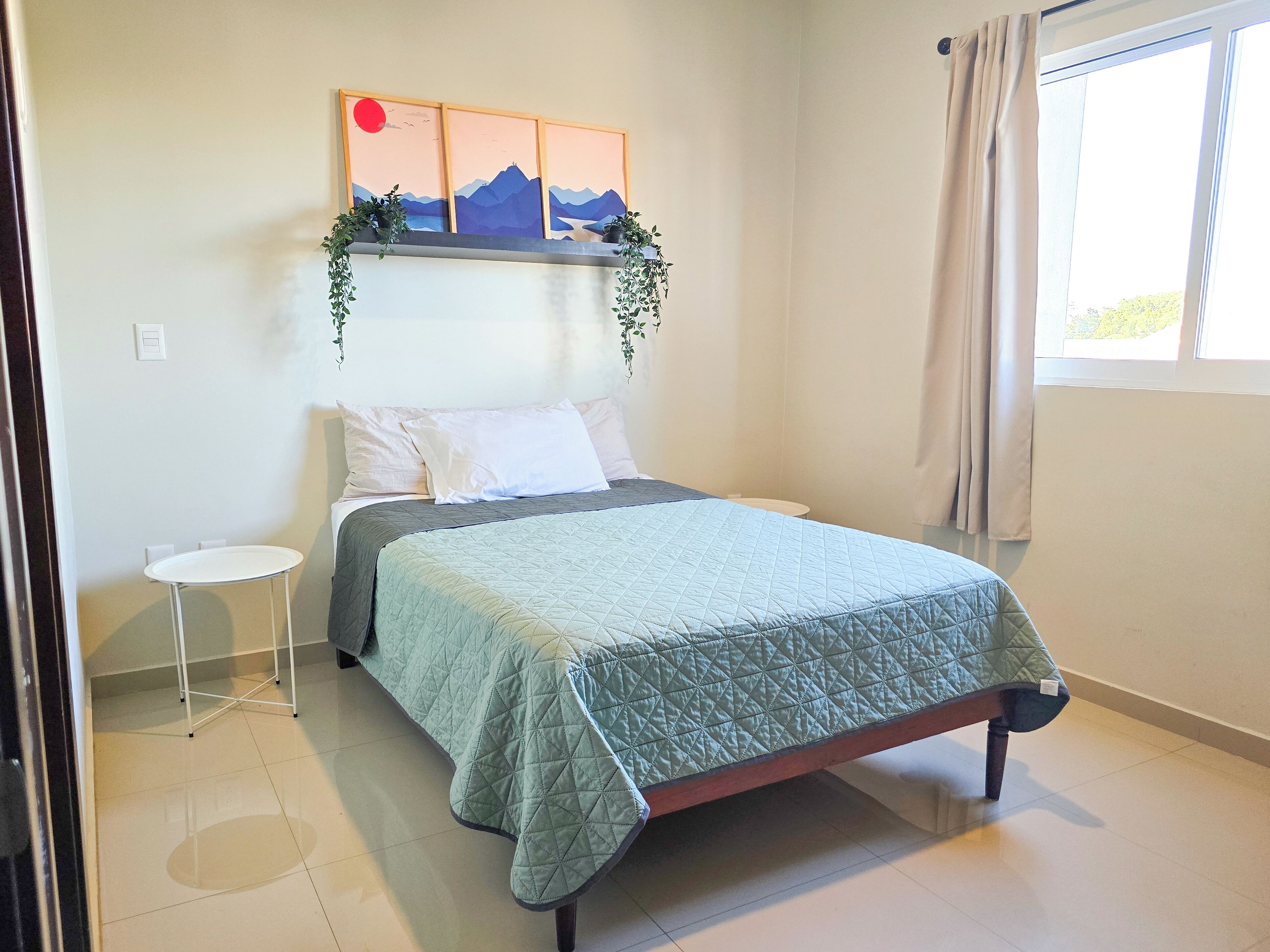 Une chambre à coucher avec un lit, une petite table de chevet, une étagère fixée au mur avec des plantes et une fenêtre avec des rideaux.