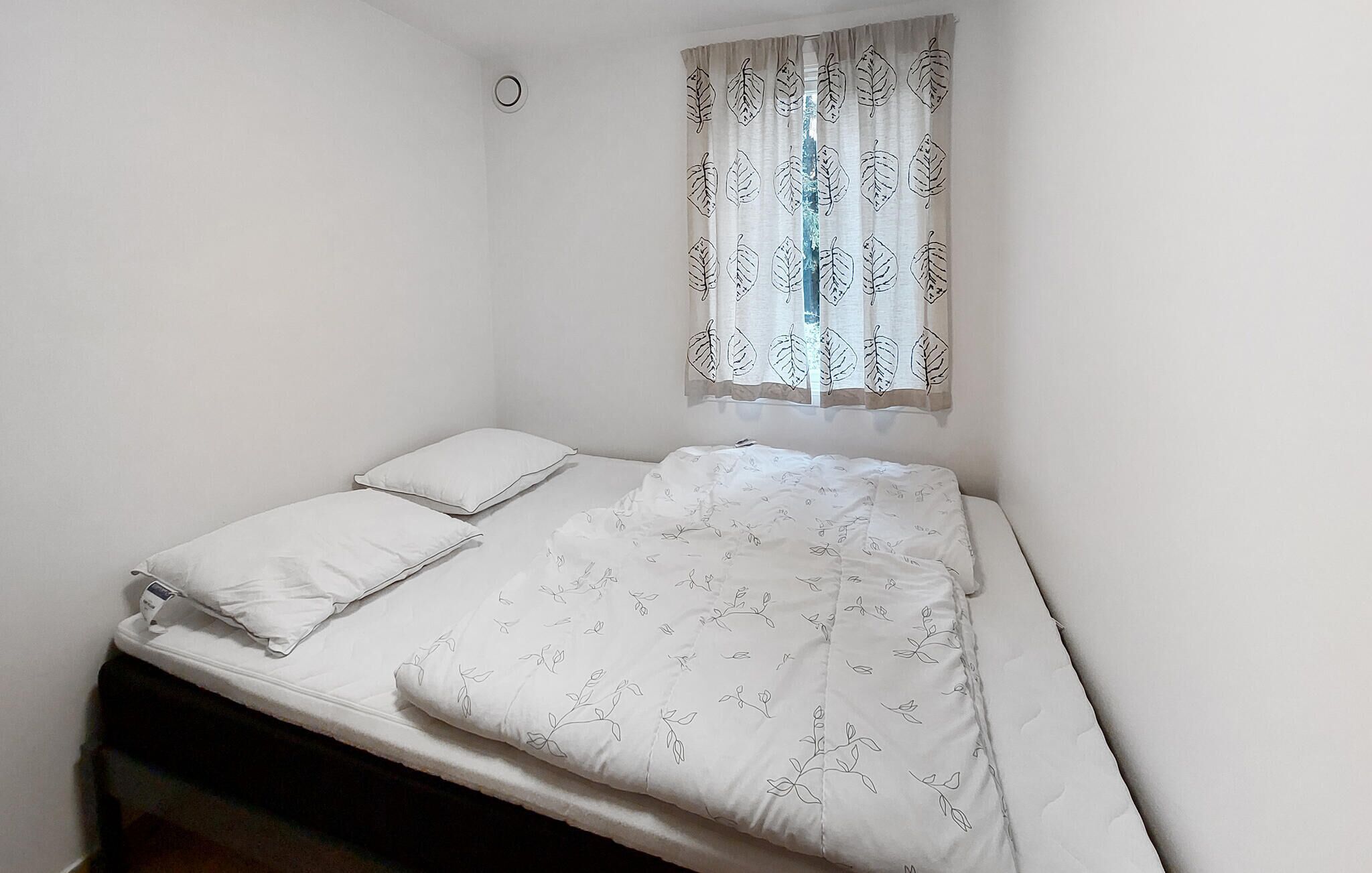 A single bed with white bedding and a floral pattern, a window with leaf-patterned curtains, and a small circular light fixture on the wall.