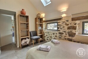 A room with a bed, a chair, wooden shelves, a stone wall, and a window.