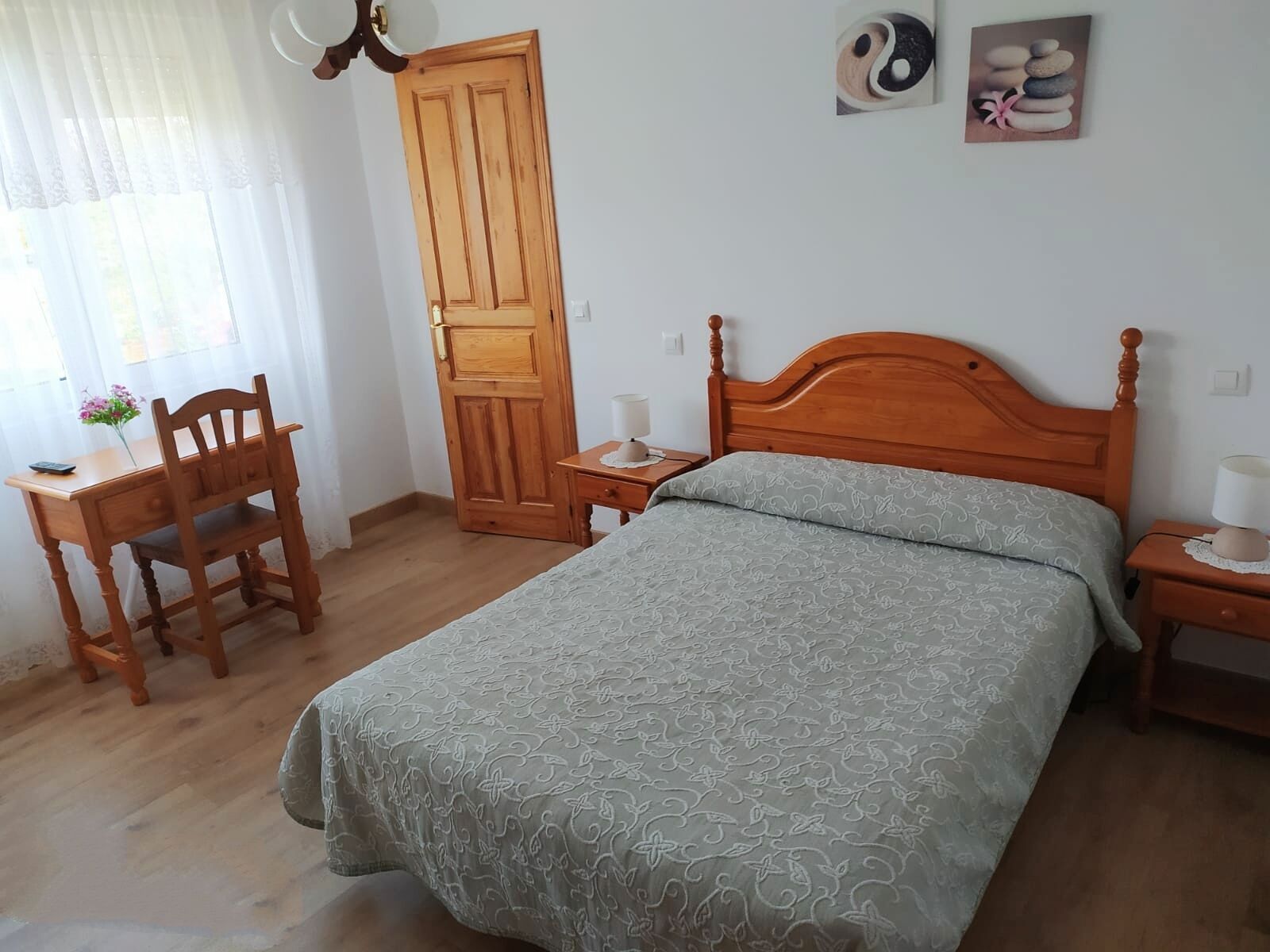 Un dormitori amb llit de fusta, escriptori de fusta amb cadira, tauleta de nit amb làmpada i una porta.