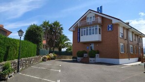 A two-story hotel with a brick facade, a balcony on the upper floor, and a parking lot in front. - Alto Santiago (San Vicente de la Barquera)