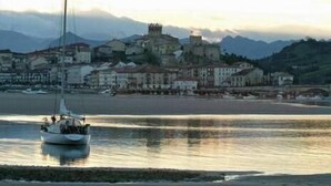 A coastal town with a prominent castle-like building, boats on the water, and a hilly backdrop. - Alto Santiago (San Vicente de la Barquera)