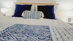 Una cama bien hecha con una colcha azul y blanca estampada, dos almohadas azul oscuro y una almohada blanca con diseño geométrico.