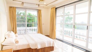 Un dormitorio con una cama grande, cortinas color beige y vista al follaje por la ventana.