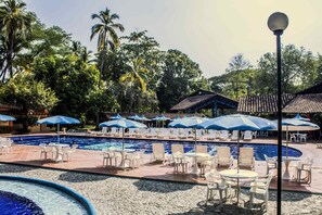 Un espace piscine doté de chaises longues et de parasols, entouré de palmiers et bordé d’un bâtiment au toit recouvert de tuiles.