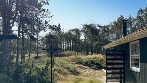 Une cabane avec une façade en bois, une fenêtre et une porte. On aperçoit une structure métallique au premier plan et une forêt en arrière-plan.