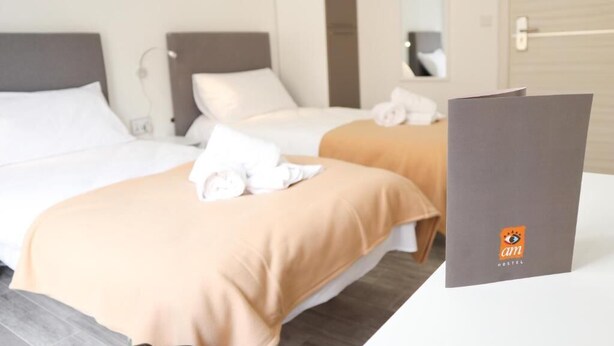 A hotel room with two beds, each with white linens and a beige blanket. There is a gray folder with the logo 'am HOSTEL' on the table. - AM Hostel (Sliema)