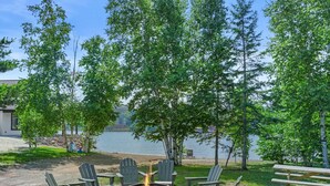 An outdoor seating area with Adirondack chairs and a fire pit, overlooking a lake with trees and a building in the background. - Waterfront Log Cabin • Sauna • Game Room • Pontoon & Snowmobile Access (Hancock)