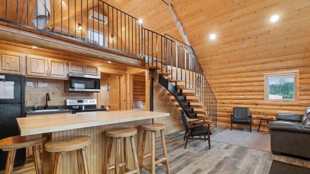 A log cabin interior with a kitchen, bar stools, a staircase, and a seating area. - Waterfront Log Cabin • Sauna • Game Room • Pontoon & Snowmobile Access (Hancock)