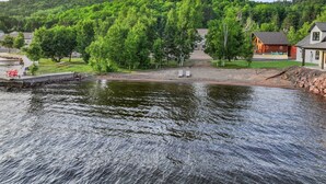 A lakeside view with a wooden dock, a small building, and a hill in the background. - Waterfront Log Cabin • Sauna • Game Room • Pontoon & Snowmobile Access (Hancock)