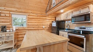 A log cabin kitchen with wooden walls, a large island, stainless steel appliances, and a window overlooking greenery. - Waterfront Log Cabin • Sauna • Game Room • Pontoon & Snowmobile Access (Hancock)