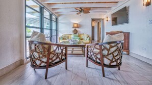 Una habitación con ventilador de techo, un sofá con cojines estampados, una mesa de centro de vidrio y sillas de madera.