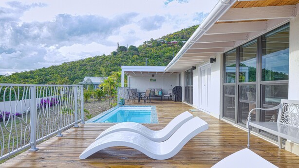 Una terraza de madera con piscina, sillas de descanso blancas y vista a una colina.