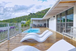 A wooden deck with a swimming pool, white lounge chairs, and a view of a hillside. - Searenity Villa in Cruz Bay with private pool and sunset view  (Cruz Bay)