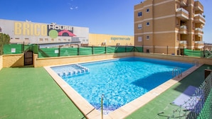 A swimming pool with a tiled deck, surrounded by a green fence. In the background, there is a building with a sign that reads "BRICO HOUSE" and another sign that says "supermercado". - Gorgeous apartment in Torrevieja (Torrevieja)