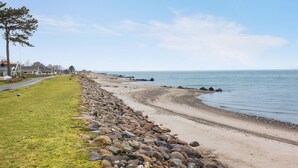 A coastal road with a stone wall leading to a beach, a grassy area, and houses in the background.