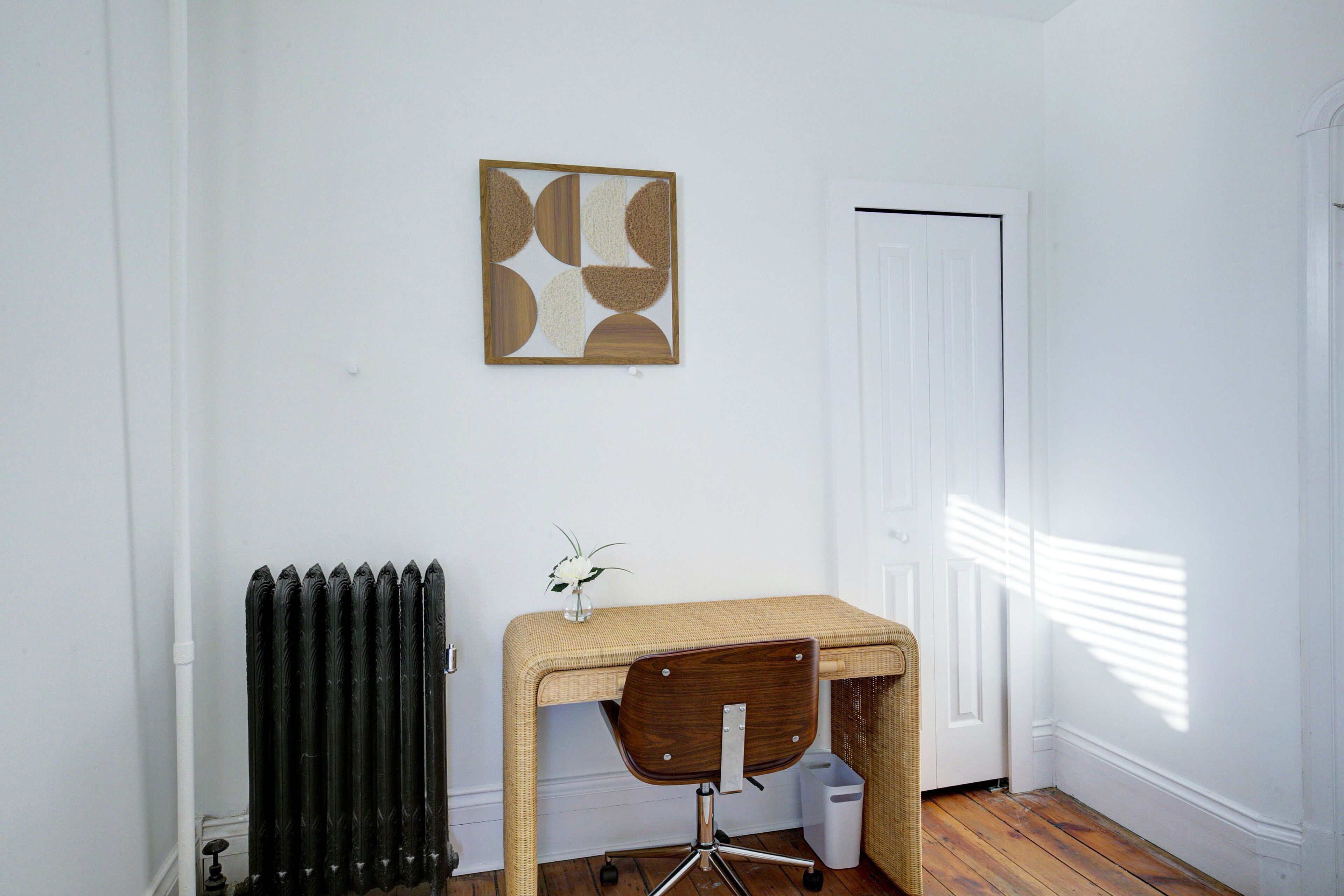 Een kamer met houten vloer, een radiator, een rieten bureau met stoel, een witte deur en een abstract kunstwerk aan de muur.