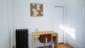 Een kamer met houten vloer, een radiator, een rieten bureau met stoel, een witte deur en een abstract kunstwerk aan de muur.