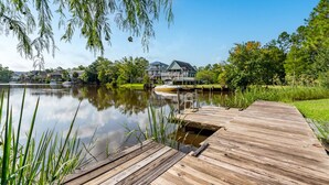 Een houten steiger die uitsteekt in een kalm meer, omgeven door weelderige begroeiing en een groot huis met een groen dak.