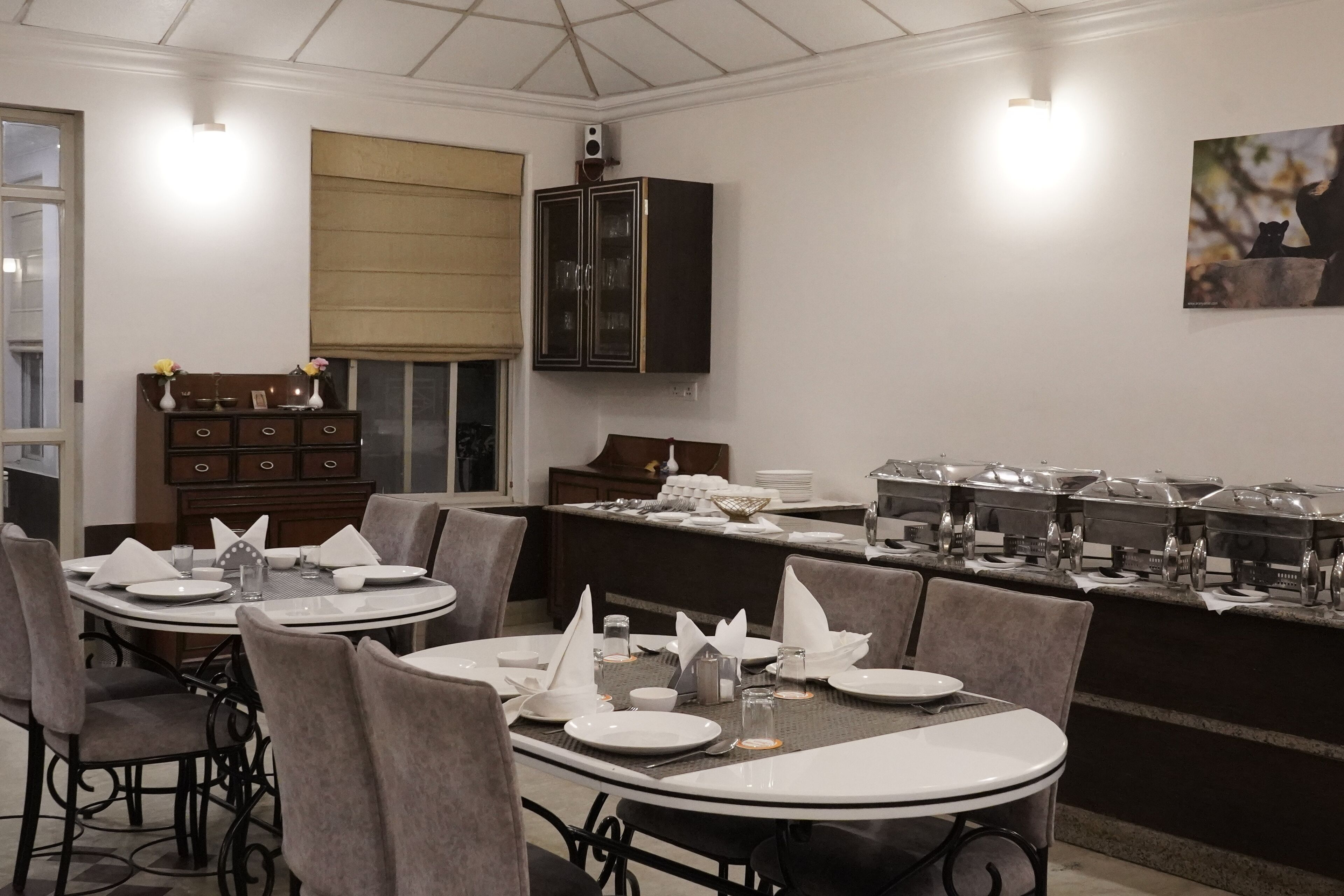 A dining area with round tables set for four, each with white plates, cutlery, and napkins. There are dark wooden cabinets and a buffet line with food containers.