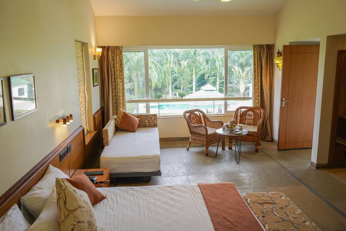 A hotel room with two beds, a dining area with wicker chairs, a view of a pool, and a wardrobe.