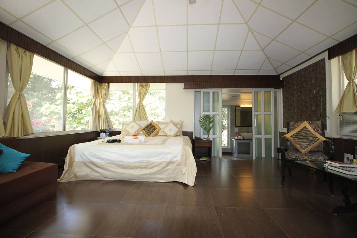 A spacious bedroom with a large bed, a sofa, and a chair. There are curtains on the windows, and a door leading to another room.