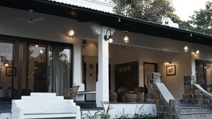 A building with a white exterior, large windows, and a stone staircase leading to a porch with wicker furniture. - Village Machaan Resort (Kurai)