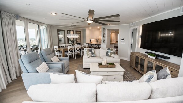 Un salon moderne doté d’un ventilateur de plafond, d’un grand téléviseur à écran plat et d’un coin repas avec des meubles blancs.