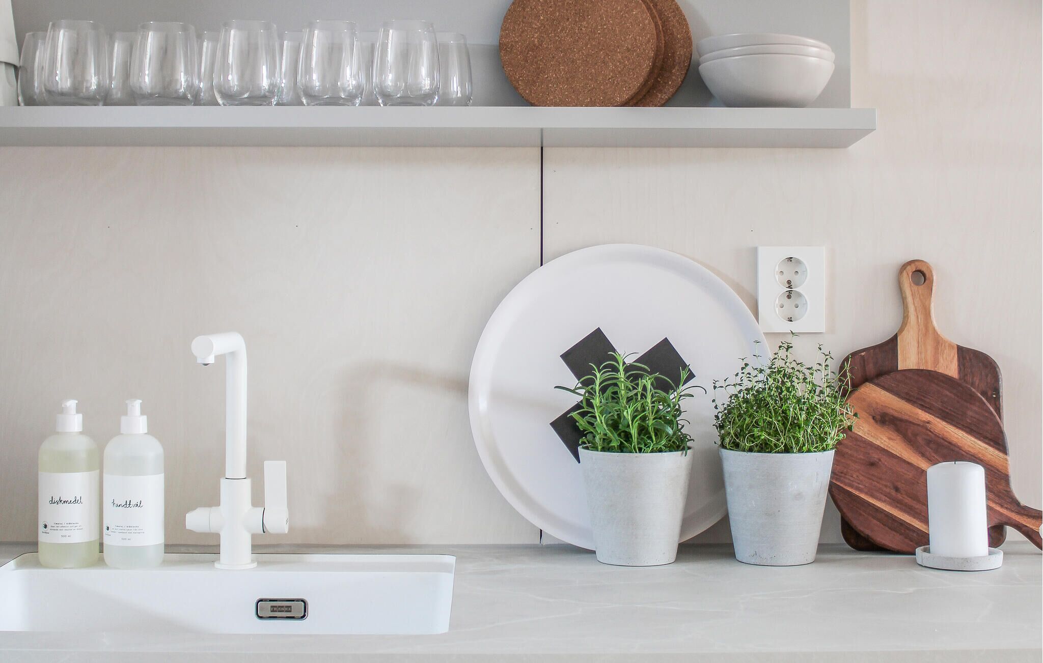 A kitchen counter with a sink, two bottles of hand soap, a white faucet, a plate with a black design, two potted plants, a wooden cutting board, and a white candle.