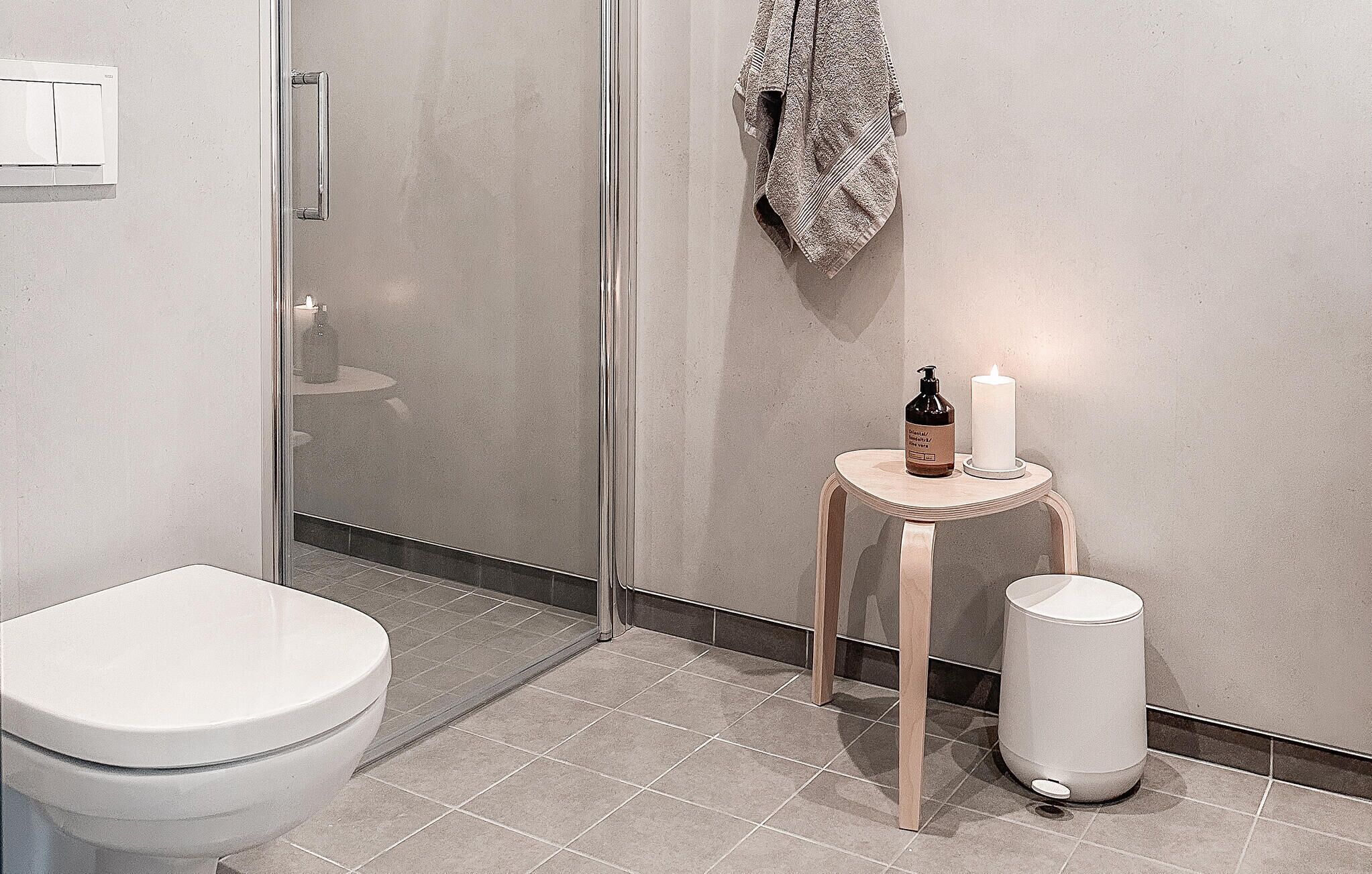 A modern bathroom with a toilet, a glass shower enclosure, a small wooden stool with a bottle and candle, and a trash bin.