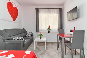 Une pièce moderne avec un canapé gris, une petite table blanche, une table rouge, une chaise grise, un téléviseur fixé au mur et une fenêtre avec des rideaux.