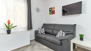 Un sofá gris con cojines estampados, un televisor de pantalla plana, una mesita, una planta en maceta y un reloj de pared.