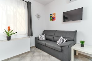 A grey sofa with patterned cushions, a flat-screen TV, a small table, a potted plant, and a wall clock.