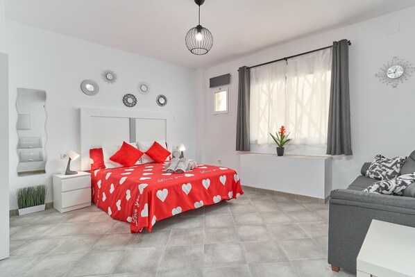 A bedroom with a bed, a nightstand, a bedside table, a window with curtains, a sofa, and a wall clock.