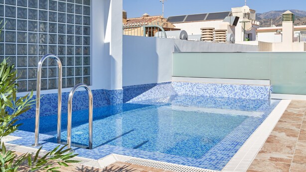 Una piscina en la azotea con baldosas azul claro, una escalera metálica y vistas de edificios circundantes.