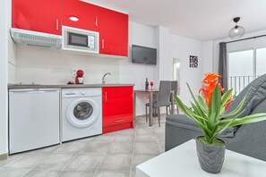 Une cuisine de taille réduite aux couleurs rouge et blanche, équipée d’un lave-linge, d’un four à micro-ondes et d’un coin repas avec un canapé gris et une plante en pot.